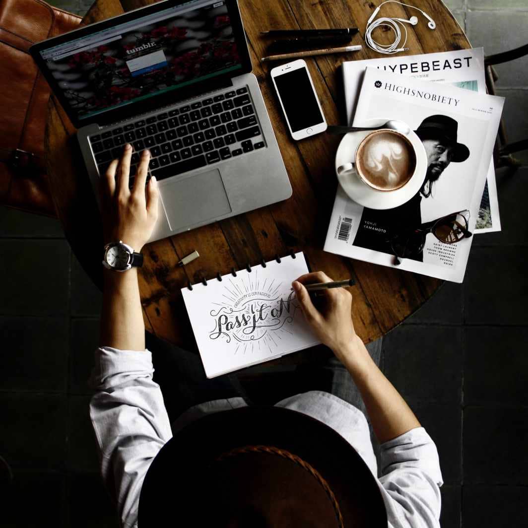 Top view of a person outsourcing graphic design in a laptop, notebook, magazine, and coffee. -